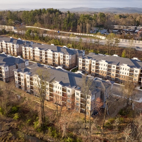 Aerial view of a modern apartment community bordered by a creek, surrounded by trees, and set against a scenic mountain backdrop